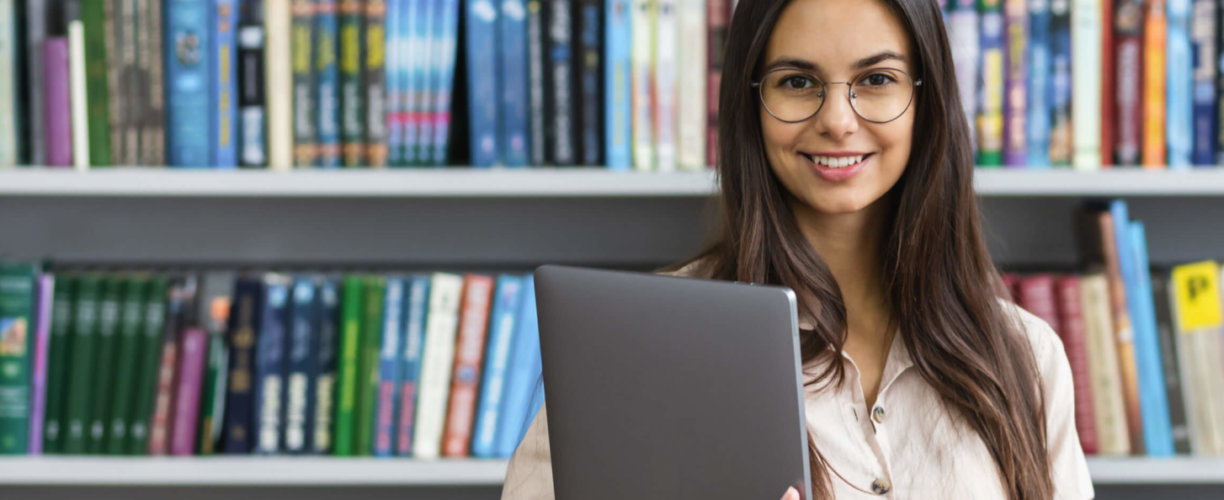 Student standing in library, holding a laptop