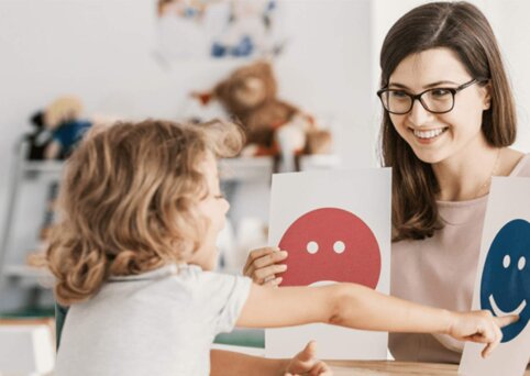 Woman sits at desk holding two paper cards with an image on each. A young child sits in front of her and is pointing at one and speaking.