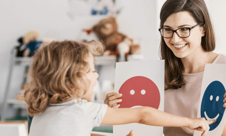 Woman sits at desk holding two paper cards with an image on each. A young child sits in front of her and is pointing at one and speaking.