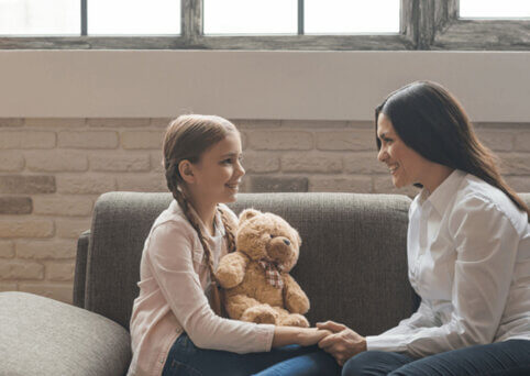 A woman on a couch sits smiles at a young girl also on the couch, holding a teddy bear. She holds her hand as they face each other.