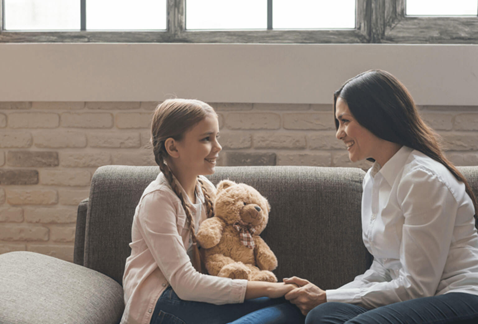 A woman on a couch sits smiles at a young girl also on the couch, holding a teddy bear. She holds her hand as they face each other.
