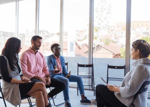 A group of people are sitting in chairs in front of a person with a clipboard. There are empty seats. They are inside and next to a wall of large glass concertina doors.