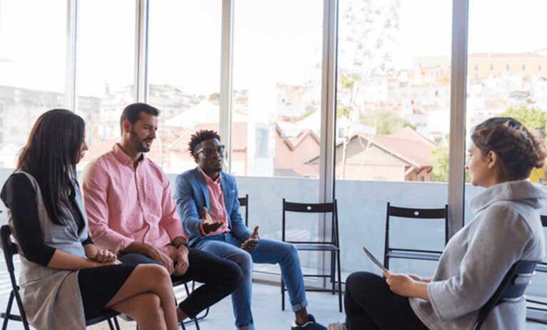 A group of people are sitting in chairs in front of a person with a clipboard. There are empty seats. They are inside and next to a wall of large glass concertina doors.