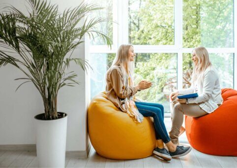 Two people sit on bean bags in front of large wall of windows. They smile at each other. One person holds a clipboard and pen. A large potted plant is in the foreground and to the left of people.