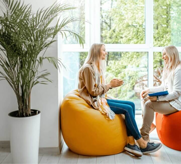 Two people sit on bean bags in front of large wall of windows. They smile at each other. One person holds a clipboard and pen. A large potted plant is in the foreground and to the left of people.