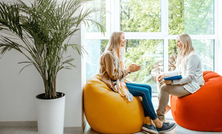 Two people sit on bean bags in front of large wall of windows. They smile at each other. One person holds a clipboard and pen. A large potted plant is in the foreground and to the left of people.