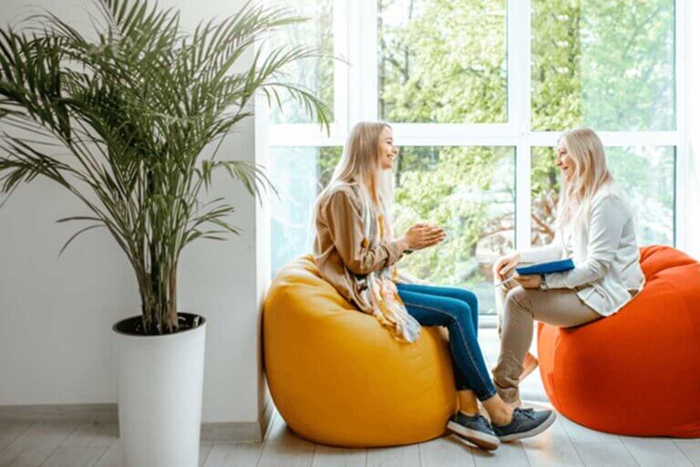 Two people sit on bean bags in front of large wall of windows. They smile at each other. One person holds a clipboard and pen. A large potted plant is in the foreground and to the left of people.