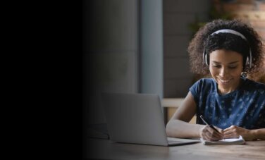 Curly haired woman wears wireless headphones and looks down at notepad she is writing on with a pen. She smiles. A laptop and glass of water on desk to the side and front of her. She appears to be in a kitchen - working at the table. A heat mat and food jars of pasta blurry in background.