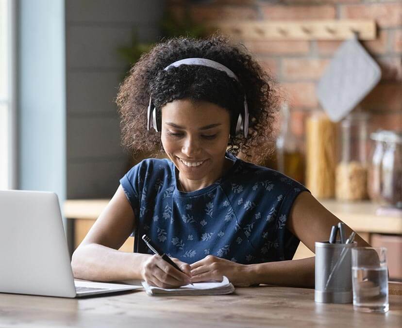 Woman with curly hair wears headphones and looks down at notepad with pen in hand. She is smiling. A laptop, pen holder and glass of water on desk in front of her. She seems to be in a kitchen - food jars of pasta blurry in background.