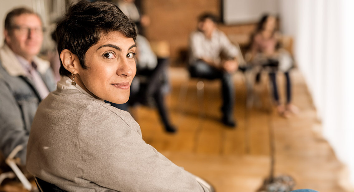 Woman with short cropped hair and padlock earring looks over her shoulder to camera. The background is a group of people in circle of chairs. They are blurry and non descript.