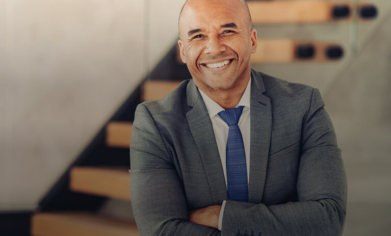 A man with a shaved head smiling towards camera, wearing a dark grey suit and blue tie - ACAP Graduate Certificate of Professional Accounting (828x676)