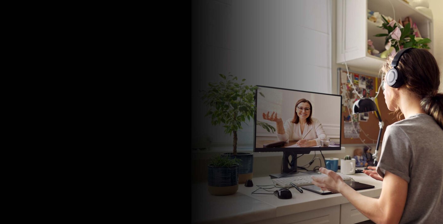 A virtual video call with a woman on a monitor screen. The monitor is on an office desk, in a home, with another person sitting in office chair with headphones. Bachelor of Psychological Science & Counselling