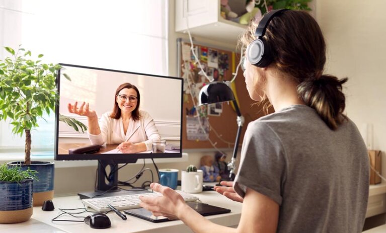 A virtual video call with a woman on a monitor screen. The monitor is on an office desk, in a home, with another person sitting in office chair with headphones. Bachelor of Psychological Science & Counselling