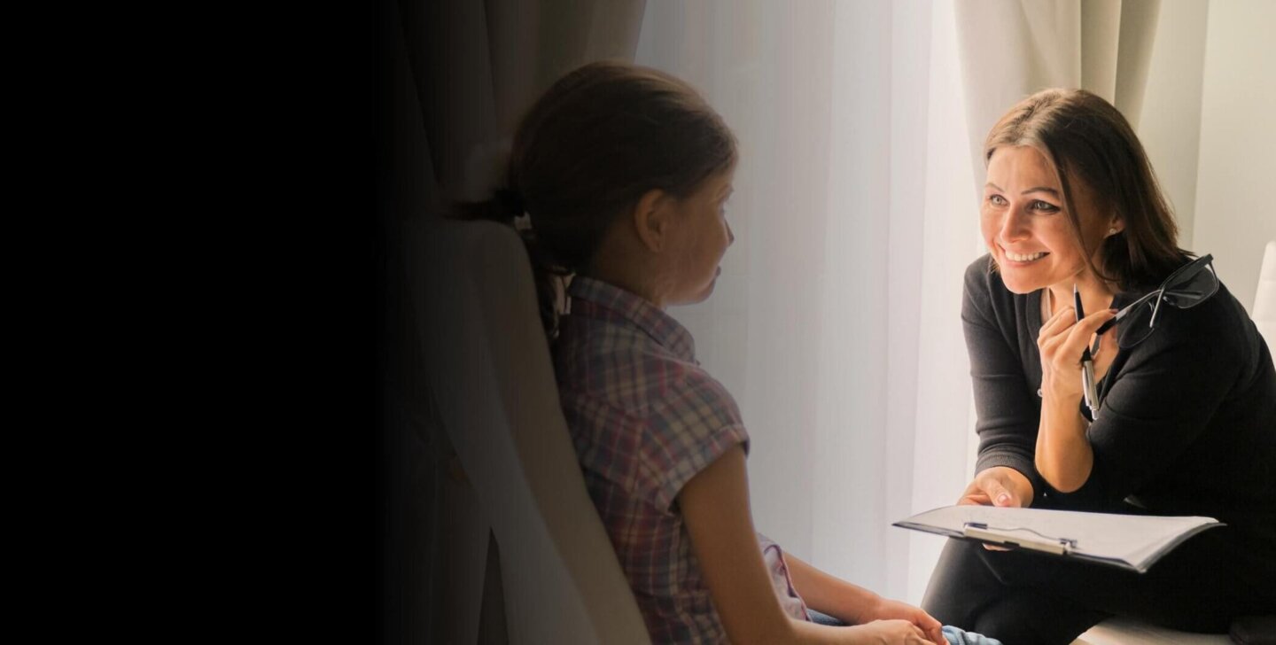 A woman sitting on a chair with a clipboard in one hand and a pen and reading glasses in the other hand, leaning towards a young child sitting in a chair opposite her | Bachelor of Social Work