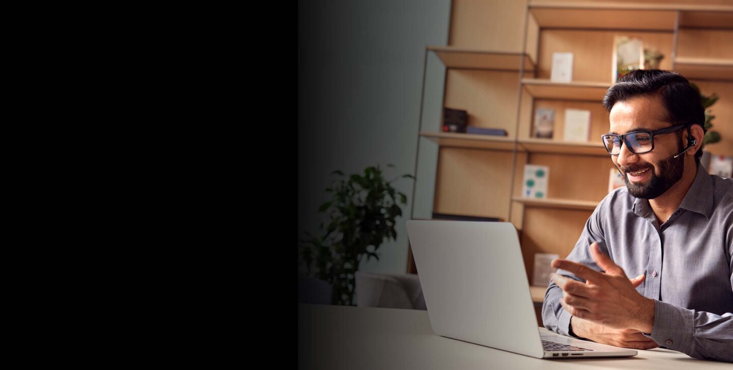 Man wears headset and sits in front of open laptop at desk. A book shelf in background