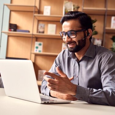 Man wears headset and sits in front of open laptop at desk. A book shelf in background