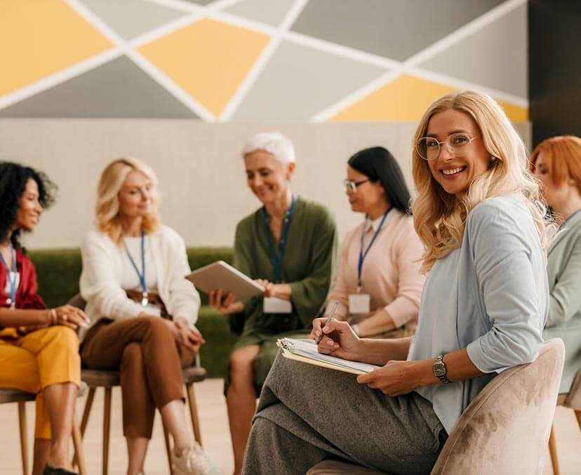 A group of woman sitting on chairs in a semicircle, with a young smiling woman turning to face camera in the foreground