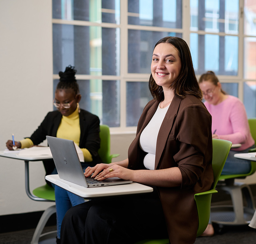 A female student working on a laptop in a classroom and smiling to camera, with two more female students working at desks in the background