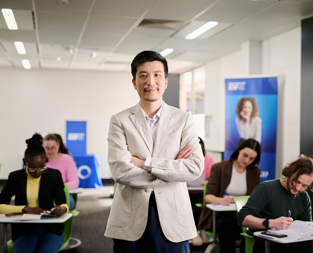 A man with short hair wearing a beige suit jacket, standing with his arms folded in a classroom with ACAP University College students.