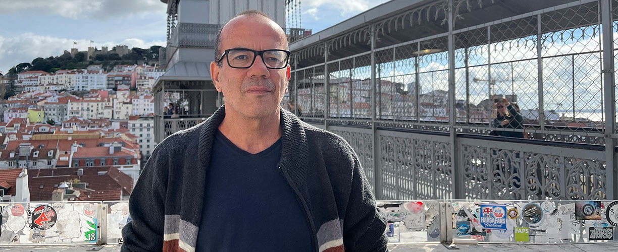 A man stands in front of walkway in a densely populated city. Posters line the walkway and apartments are seen covering the hillside behind.