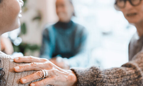 Woman outstretches hand which rests on another persons shoulder. The person is seen in profile and is wearing earrings and facing person with hand on their shoulder.