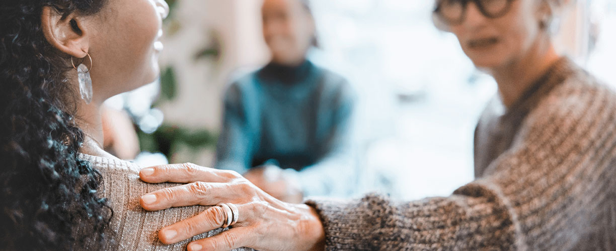 Woman outstretches hand which rests on another persons shoulder. The person is seen in profile and is wearing earrings and facing person with hand on their shoulder.