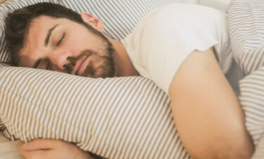 Man with a beard and wearing a white tee shirt - rests with head on pillow and doona covering him. He is lying in bed and the bedhead is just visible in background.