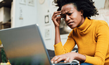 Person wearing bright turtleneck sits on couch in a home. The laptop is on a table in front of them. They squint and frown at screen with hand to forehead.