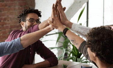Group of four people sit around a table. Each one are raising one arm - there hands meet in the centre of table - high in the air. High five pose with four work colleagues.