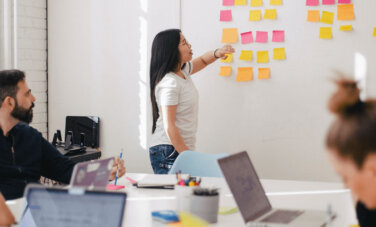 Stand up meeting with post it notes on a while - agile methodologies. Woman is speaking to wall of post it notes. People sit around a boardroom table, laptops and pens in hand - also looking at wall.