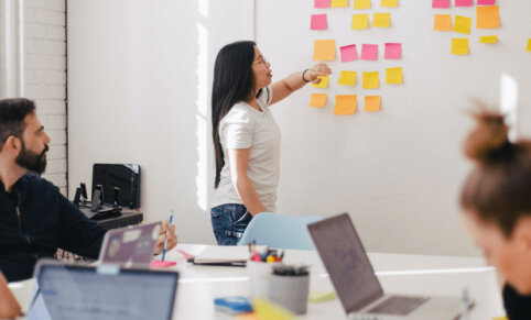 Stand up meeting with post it notes on a while - agile methodologies. Woman is speaking to wall of post it notes. People sit around a boardroom table, laptops and pens in hand - also looking at wall.