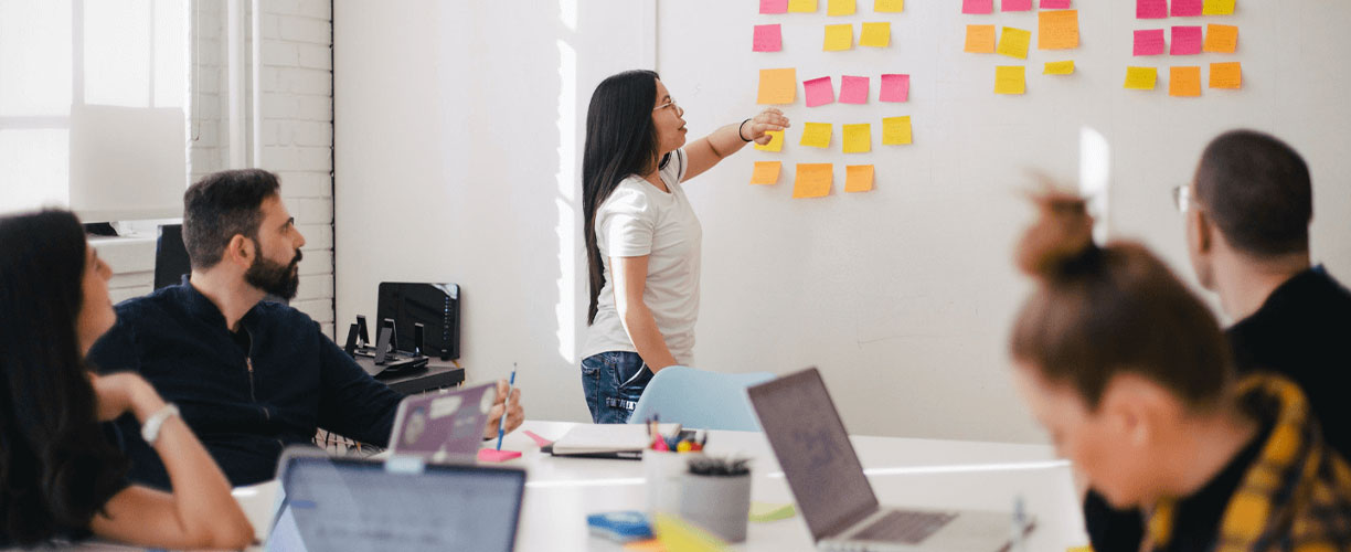 Stand up meeting with post it notes on a while - agile methodologies. Woman is speaking to wall of post it notes. People sit around a boardroom table, laptops and pens in hand - also looking at wall.