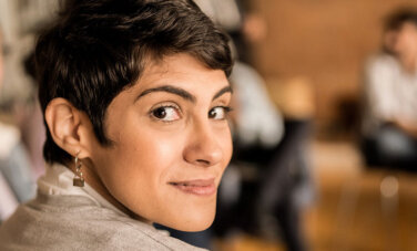 Woman with cropped hair looks over her shoulder and at camera. Group of people in background appear to be sitting in a circle on chairs.