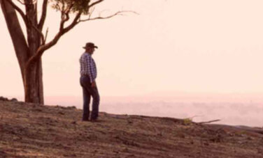 Single figure of man on hill side looking out. One lone tree in background. Little to no grass or trees on the land. Dry. Arid. Man wears a checkered collared shirt and old farming hat