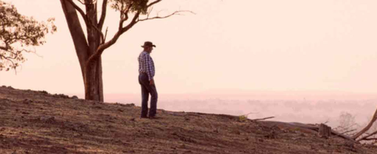 Single figure of man on hill side looking out. One lone tree in background. Little to no grass or trees on the land. Dry. Arid. Man wears a checkered collared shirt and old farming hat
