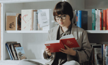 Woman sits cross legged on the floor in library. Books on shelf behind them.