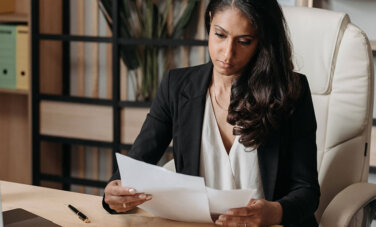 Woman sitting at desk in an office chair. She is wearing a suit. A laptop and a closed pen on desk in front of her. She is looking at the papers in her hand. A wall of shelves are behind her.