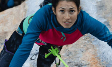 Woman climbs on rockface. She is wearing a harness, a large green rope is tied to her waist and carabiners are dangling from her belt.