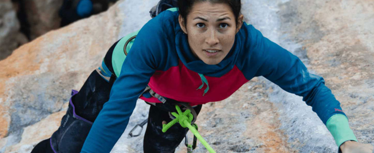 Woman climbs on rockface. She is wearing a harness, a large green rope is tied to her waist and carabiners are dangling from her belt.