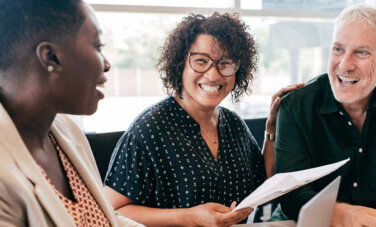 Three people sit at table. One person with cropped hair, a curly haired person and a white haired person. They all smile at each other and sit at a table.