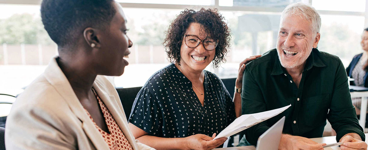 Three people sit at table. One person with cropped hair, a curly haired person and a white haired person. They all smile at each other and sit at a table.
