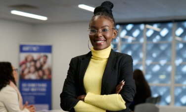 Woman in a suit stands in a room with people in the background. She smiles at the camera with arms crossed.