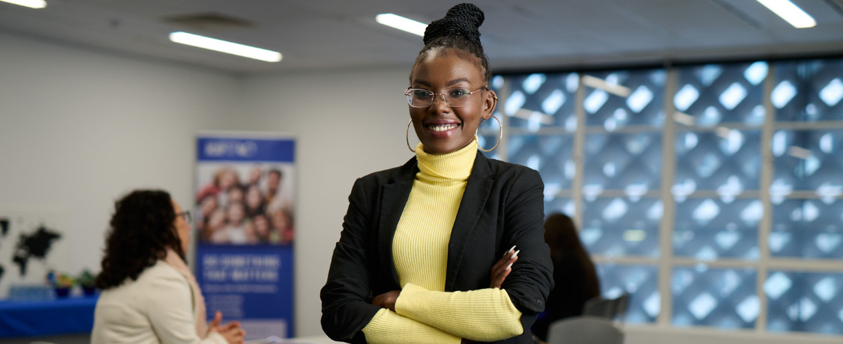 Woman in a suit stands in a room with people in the background. She smiles at the camera with arms crossed.