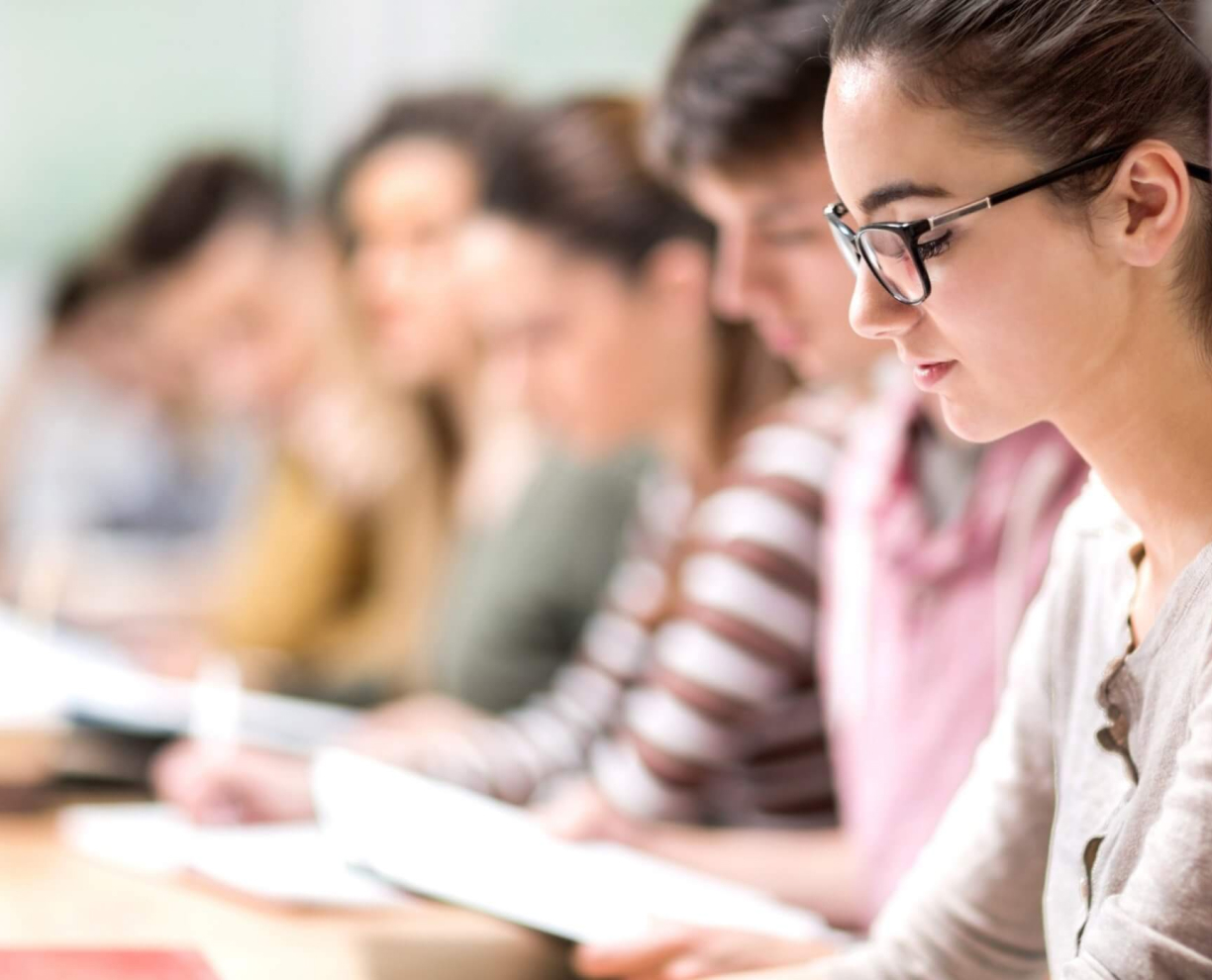Woman looks down at desk. Multiple people are in the background - blurred but looking down at desk. Image is of students in a seated row or desk.