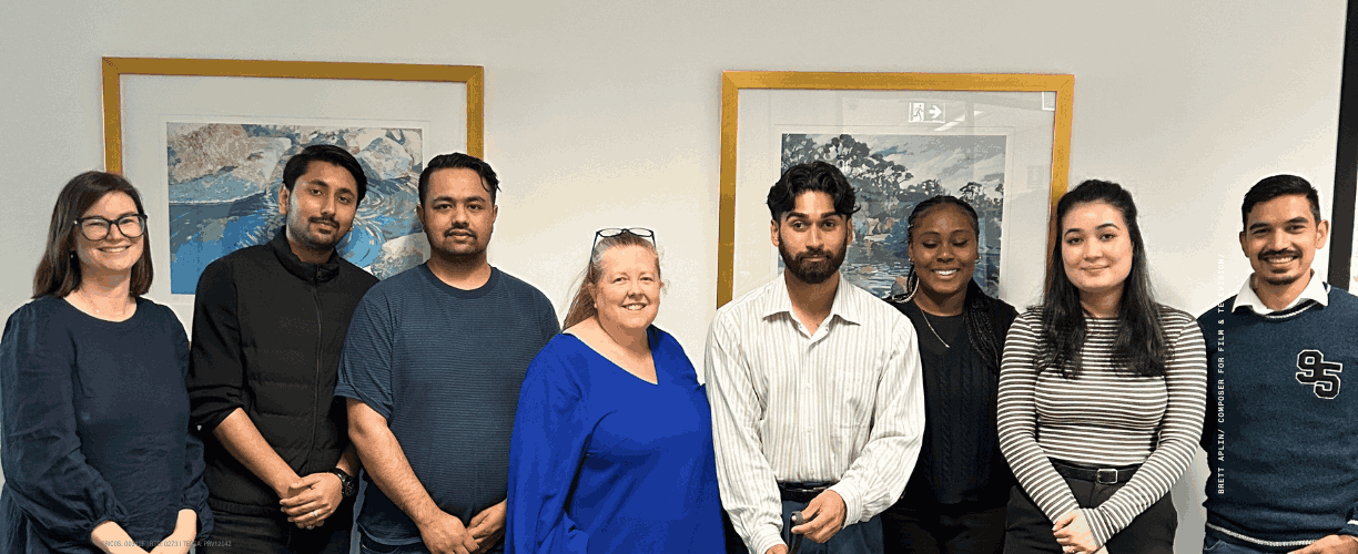 Eight people standing in front of a blank wall with two frames hanging. They are of mixed ethnicities and smiling at the camera with a dog in the foreground.