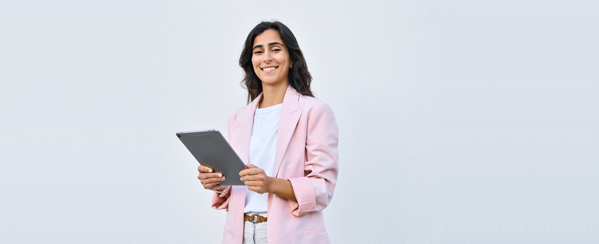 A woman wearing a pink suit jacket, standing with a tablet in hand in front of a grey background