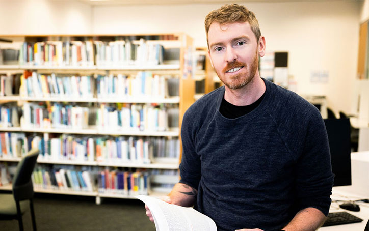 Man holding book stands in a room. Books on bookshelves on wall behind.