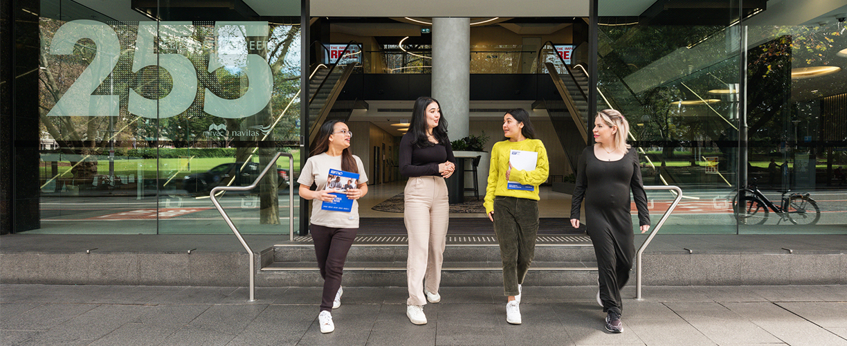 Four women walk out from office building onto city street. They all look at each other and two are holding pamphlets. Number 255 on glass front wall.