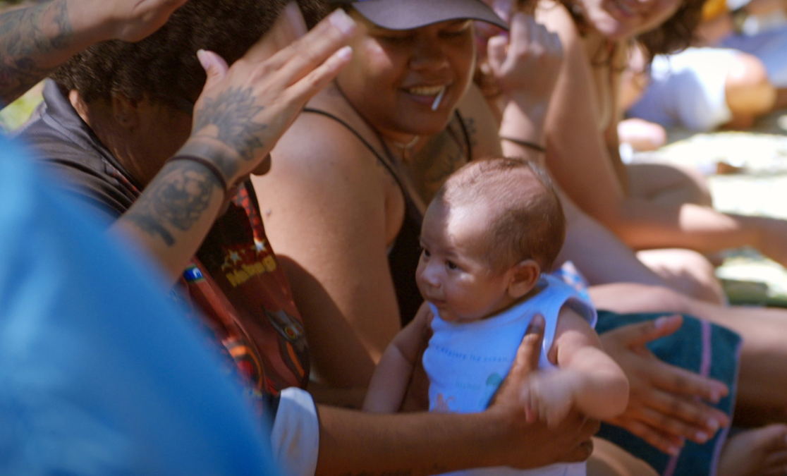 Image of baby being held in a group of people as someone raises tattooed hands above its heads
