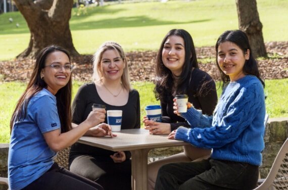 Four female high school students sitting at an outdoor table on campus with coffees in their hands - 761x761